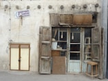 A rustic and worn storefront with wooden shutters and a weathered door. A small stool and various items are stored behind glass doors. A sign indicating a 'Holy Shrine' is mounted on the wall.