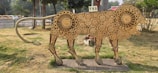 A sculpture of a lion made entirely from industrial gears of various sizes is set on a grassy area. The sculpture has a linear, stylized form and appears to be part of an outdoor display or installation. In the background, there are trees, a road with passing vehicles, and a sign that reads 'TRS.' The base of the sculpture features the text 'MAKE IN INDIA.'