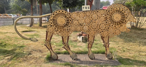 A sculpture of a lion made entirely from industrial gears of various sizes is set on a grassy area. The sculpture has a linear, stylized form and appears to be part of an outdoor display or installation. In the background, there are trees, a road with passing vehicles, and a sign that reads 'TRS.' The base of the sculpture features the text 'MAKE IN INDIA.'