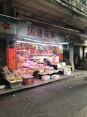 A street-side fruit stall is displaying a wide variety of colorful fruits in baskets and crates. A person is tending to the stall, organizing or selling produce. Signs with Chinese characters adorn the shop, and there are decorative red ornaments hanging above the fruits.