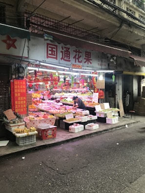 A street-side fruit stall is displaying a wide variety of colorful fruits in baskets and crates. A person is tending to the stall, organizing or selling produce. Signs with Chinese characters adorn the shop, and there are decorative red ornaments hanging above the fruits.