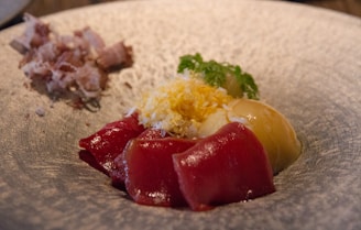A dish featuring vibrant red tuna slices, complemented by a creamy-looking sauce and garnished with finely grated cheese or egg. Fresh green herbs add a touch of color and freshness. On the side, there appears to be a portion of shredded meat or fish.
