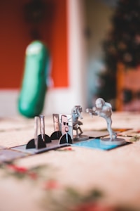 Several miniature figures set up on a board game with a background featuring a vertically shaped plush that resembles a pickle. The figures seem to be engaged in a scenario featuring characters with weapons posed as if in combat. The environment features a soft focus with warm lighting giving it a cozy, indoor feel.