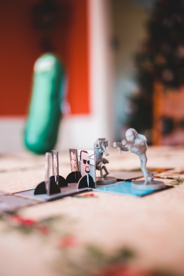 Several miniature figures set up on a board game with a background featuring a vertically shaped plush that resembles a pickle. The figures seem to be engaged in a scenario featuring characters with weapons posed as if in combat. The environment features a soft focus with warm lighting giving it a cozy, indoor feel.