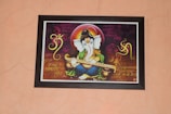A framed artwork of a Hindu deity with an elephant head, holding a musical instrument. The background features Sanskrit or Hindi script in ornate calligraphy, along with decorative patterns and symbols, including an 'Om' symbol. The figure is adorned in bright, vibrant colors and ornamentation.