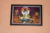 A framed artwork of a Hindu deity with an elephant head, holding a musical instrument. The background features Sanskrit or Hindi script in ornate calligraphy, along with decorative patterns and symbols, including an 'Om' symbol. The figure is adorned in bright, vibrant colors and ornamentation.