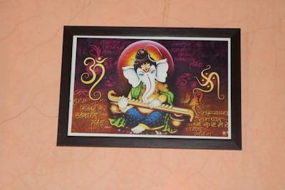 A framed artwork of a Hindu deity with an elephant head, holding a musical instrument. The background features Sanskrit or Hindi script in ornate calligraphy, along with decorative patterns and symbols, including an 'Om' symbol. The figure is adorned in bright, vibrant colors and ornamentation.