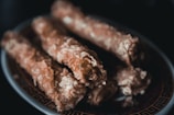 Several deep-fried, batter-coated rolls or sticks are arranged on a detailed patterned plate. The coating appears crispy and slightly oily, with a golden brown hue.