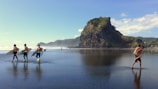 A group of surfers carrying surfboards walks across a wet sandy beach reflecting the blue sky, with a prominent rocky outcrop and lush green hillside in the background. Another man carries someone on his back while walking through the shallow water.