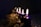 A row of lit candles with tall, flickering flames sits on a decorated surface. The candles are a deep purple color and are surrounded by decorative, shimmering gold elements. The background is dark, enhancing the warm glow of the candlelight and creating an intimate atmosphere.