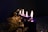 A row of lit candles with tall, flickering flames sits on a decorated surface. The candles are a deep purple color and are surrounded by decorative, shimmering gold elements. The background is dark, enhancing the warm glow of the candlelight and creating an intimate atmosphere.