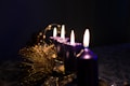 A row of lit candles with tall, flickering flames sits on a decorated surface. The candles are a deep purple color and are surrounded by decorative, shimmering gold elements. The background is dark, enhancing the warm glow of the candlelight and creating an intimate atmosphere.