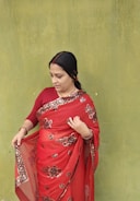 A woman wearing a red saree with floral patterns stands against a textured green wall. Her hair is tied back, and she appears to be adjusting or examining her saree. The contrast between the vibrant red saree and the muted green wall is striking.