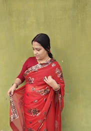 A woman wearing a red saree with floral patterns stands against a textured green wall. Her hair is tied back, and she appears to be adjusting or examining her saree. The contrast between the vibrant red saree and the muted green wall is striking.