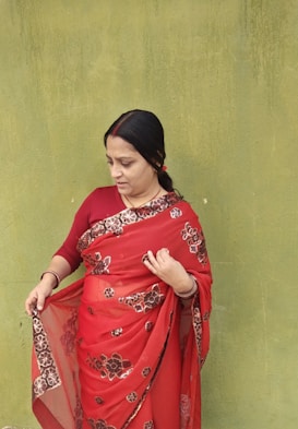 A woman wearing a red saree with floral patterns stands against a textured green wall. Her hair is tied back, and she appears to be adjusting or examining her saree. The contrast between the vibrant red saree and the muted green wall is striking.