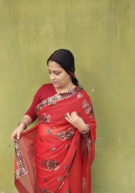 A woman wearing a red saree with floral patterns stands against a textured green wall. Her hair is tied back, and she appears to be adjusting or examining her saree. The contrast between the vibrant red saree and the muted green wall is striking.