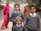 Three children dressed in school uniforms stand in a rural setting. The two girls and one boy wear matching grey sweaters and ties. Behind them, two women, one in a pink top and the other wrapped in a brown shawl, are slightly out of focus, standing near a road. The background features a natural landscape with a tree and an unclear road leading into the distance.