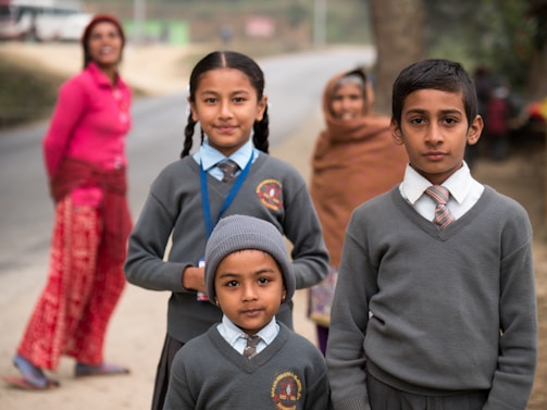 Three children dressed in school uniforms stand in a rural setting. The two girls and one boy wear matching grey sweaters and ties. Behind them, two women, one in a pink top and the other wrapped in a brown shawl, are slightly out of focus, standing near a road. The background features a natural landscape with a tree and an unclear road leading into the distance.