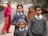 Three children dressed in school uniforms stand in a rural setting. The two girls and one boy wear matching grey sweaters and ties. Behind them, two women, one in a pink top and the other wrapped in a brown shawl, are slightly out of focus, standing near a road. The background features a natural landscape with a tree and an unclear road leading into the distance.