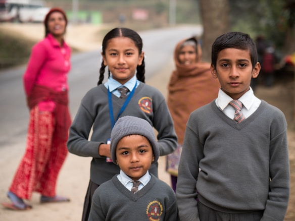 Three children dressed in school uniforms stand in a rural setting. The two girls and one boy wear matching grey sweaters and ties. Behind them, two women, one in a pink top and the other wrapped in a brown shawl, are slightly out of focus, standing near a road. The background features a natural landscape with a tree and an unclear road leading into the distance.