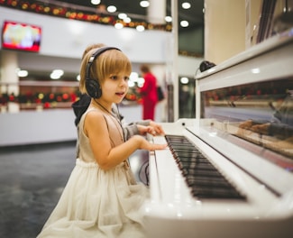 A young child wearing a sleeveless dress and headphones is playing a white piano. There are blurred figures and festive decorations in the background, indicating a possible public or festive setting. The child is focused on the piano keys.