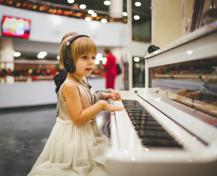 A young child wearing a sleeveless dress and headphones is playing a white piano. There are blurred figures and festive decorations in the background, indicating a possible public or festive setting. The child is focused on the piano keys.