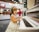 A young child wearing a sleeveless dress and headphones is playing a white piano. There are blurred figures and festive decorations in the background, indicating a possible public or festive setting. The child is focused on the piano keys.