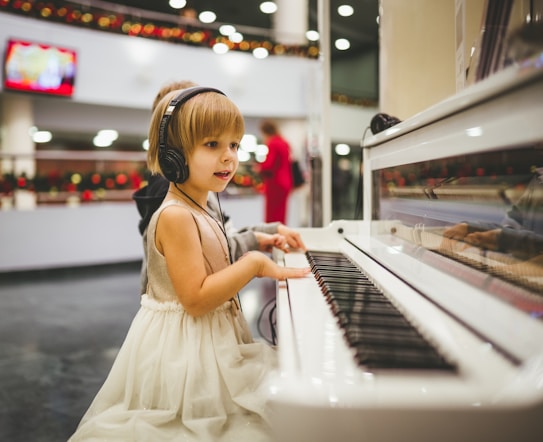 A young child wearing a sleeveless dress and headphones is playing a white piano. There are blurred figures and festive decorations in the background, indicating a possible public or festive setting. The child is focused on the piano keys.