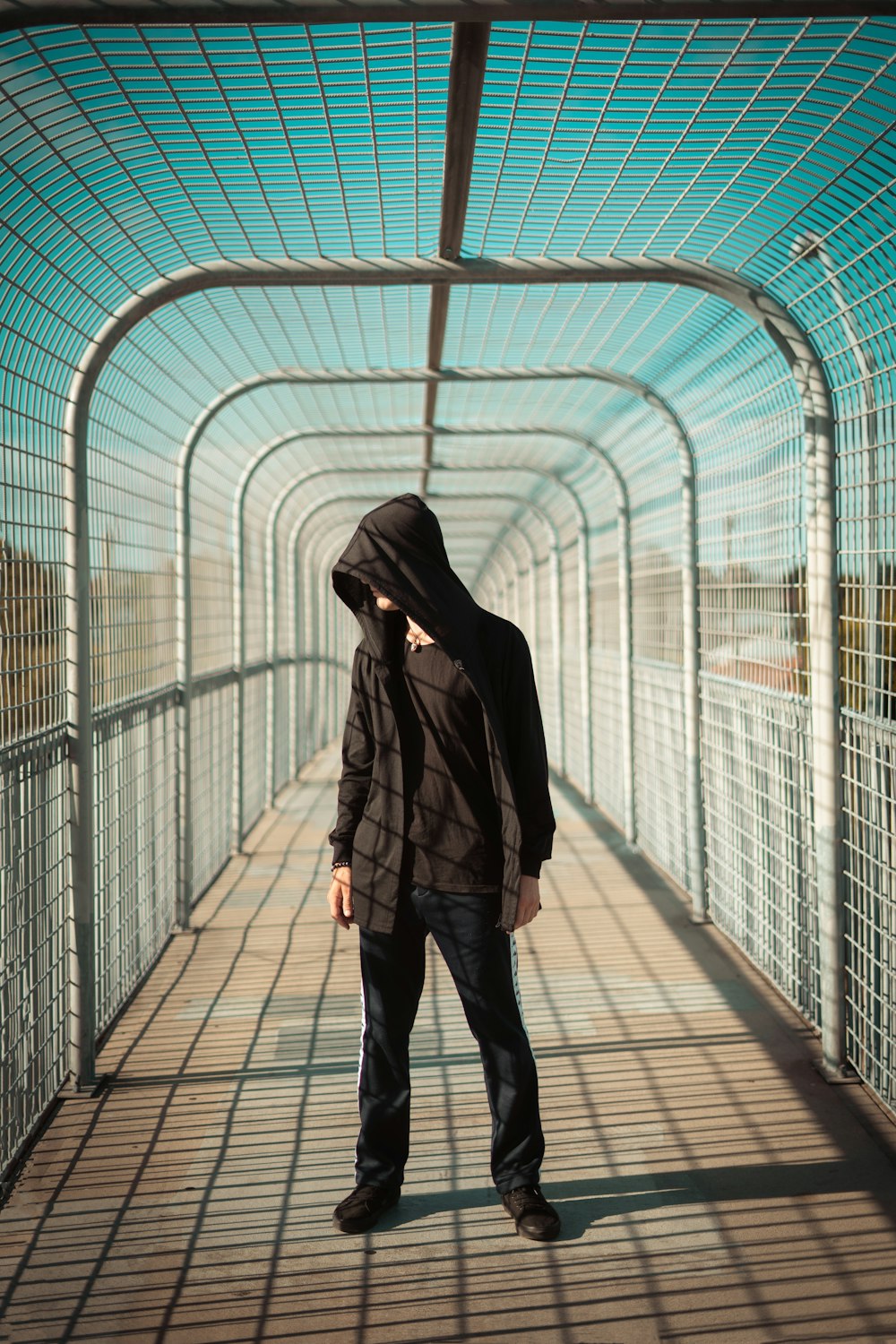 man wearing black hooded jacket standing on steel bridge under blue and white sky