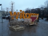 A large yellow sign with the word 'Witamy' in a public square, decorated with a lighted reindeer and sleigh display in the foreground. The background includes leafless trees and buildings under a cloudy, overcast sky.