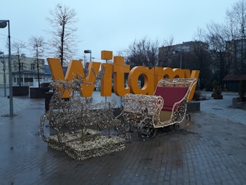 A large yellow sign with the word 'Witamy' in a public square, decorated with a lighted reindeer and sleigh display in the foreground. The background includes leafless trees and buildings under a cloudy, overcast sky.