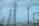 A network of tall metal electricity pylons and wires set against a cloudy blue sky. The scene includes various electrical structures and some urban environmental elements in the background.