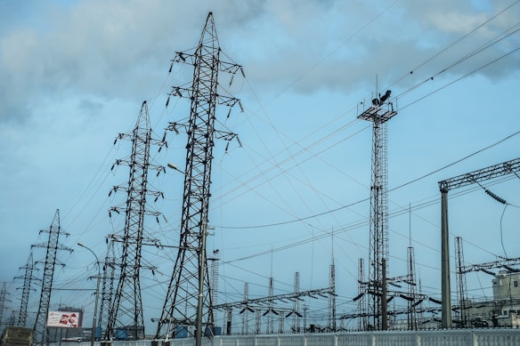 A network of tall metal electricity pylons and wires set against a cloudy blue sky. The scene includes various electrical structures and some urban environmental elements in the background.