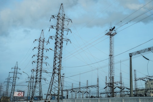 A network of tall metal electricity pylons and wires set against a cloudy blue sky. The scene includes various electrical structures and some urban environmental elements in the background.