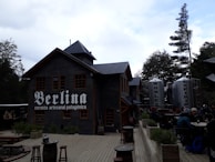 A rustic building made of wood with a sign that says 'Berlina cerveza artesanal patagónica'. Adjacent are large metallic tanks labeled 'Nobleza' and 'Espíritu'. There is outdoor seating with people gathered around tables, surrounded by trees and a wooden deck area.