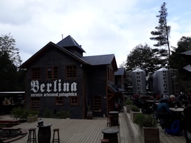 A rustic building made of wood with a sign that says 'Berlina cerveza artesanal patag&oacute;nica'. Adjacent are large metallic tanks labeled 'Nobleza' and 'Esp&iacute;ritu'. There is outdoor seating with people gathered around tables, surrounded by trees and a wooden deck area.