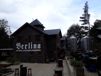 A rustic building made of wood with a sign that says 'Berlina cerveza artesanal patagónica'. Adjacent are large metallic tanks labeled 'Nobleza' and 'Espíritu'. There is outdoor seating with people gathered around tables, surrounded by trees and a wooden deck area.