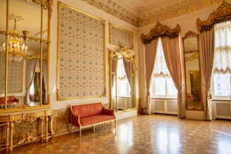 An opulent room with ornate gold-framed mirrors and richly decorated walls. A red velvet sofa and luxurious drapes contribute to the elegance. The floor features intricate woodwork patterns.