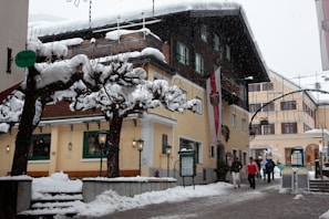 A picturesque street in a snowy European town, featuring traditional buildings with snow-covered roofs and leafless trees lining the pathway. Several people in winter clothing are walking along the street, adding a lively touch to the peaceful wintery scene. Decorative lights and signs add a quaint charm to the area.