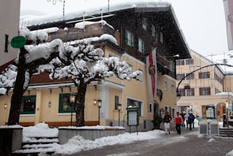 A picturesque street in a snowy European town, featuring traditional buildings with snow-covered roofs and leafless trees lining the pathway. Several people in winter clothing are walking along the street, adding a lively touch to the peaceful wintery scene. Decorative lights and signs add a quaint charm to the area.