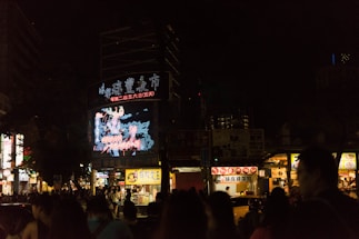A bustling city street at night with a large digital billboard displaying bright, colorful graphics in front of a crowd of people. Vendor stalls with illuminated signs line the street, offering food and goods.