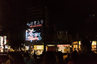 A bustling city street at night with a large digital billboard displaying bright, colorful graphics in front of a crowd of people. Vendor stalls with illuminated signs line the street, offering food and goods.