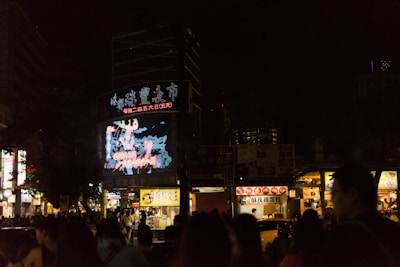 A bustling city street at night with a large digital billboard displaying bright, colorful graphics in front of a crowd of people. Vendor stalls with illuminated signs line the street, offering food and goods.