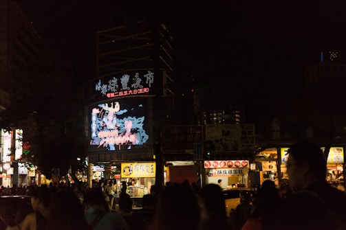 A bustling city street at night with a large digital billboard displaying bright, colorful graphics in front of a crowd of people. Vendor stalls with illuminated signs line the street, offering food and goods.
