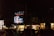 A bustling city street at night with a large digital billboard displaying bright, colorful graphics in front of a crowd of people. Vendor stalls with illuminated signs line the street, offering food and goods.