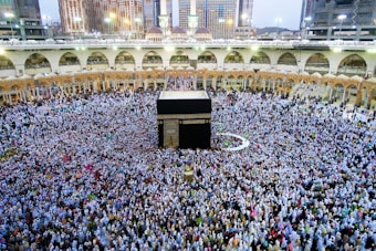 A large gathering of people surrounds a central black structure adorned with gold details. The structure is situated within an expansive courtyard encircled by multi-level arches and buildings with intricate designs. The crowd appears to be engaged in a religious practice, with many wearing modest garments in a variety of colors.