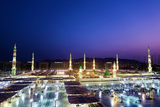An expansive view of an illuminated mosque complex is seen at twilight, with several tall minarets reaching up into a deepening blue and purple sky. The golden lighting highlights the architectural details, and a dome with a green top is visible in the foreground. Numerous people can be seen moving around in the courtyard, and the overall scene gives a sense of tranquility and reverence.