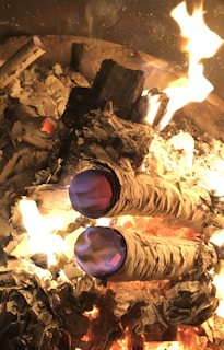 A close-up view of a fire pit containing burning logs with ashes scattered around. Two prominent bundles, likely sage or similar materials, are smoldering with vibrant blue and purple flames emerging. The fire emits a warm glow, illuminating surrounding charred wood and paper remnants.