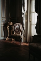 A vintage room with ornate decor, featuring an elegant, upholstered chair with floral patterns placed on a wooden floor. The room is adorned with intricate woodwork and a vintage pedestal. Soft light filters through sheer curtains, casting shadows and creating a serene ambience.