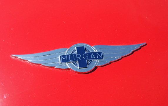A silver Morgan car emblem with wings is centered on a glossy red background. The emblem features the word 'Morgan' across a blue cross-like design.